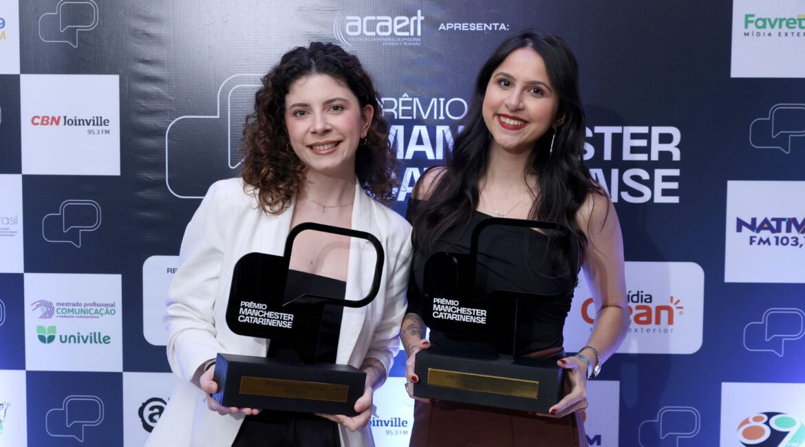 Duas mulheres sorrindo e segurando troféus do Prêmio Manchester Catarinense em frente a um painel com logotipos de patrocinadores e apoiadores do evento.