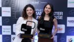Duas mulheres sorrindo e segurando troféus do Prêmio Manchester Catarinense em frente a um painel com logotipos de patrocinadores e apoiadores do evento.